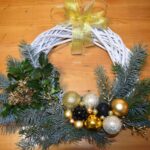 422 Biały pleciony wianek z dużą ilością gałązek świerku, złotymi bombkami i kokardą.