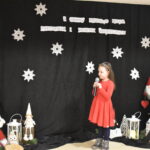 Dziewczynka w czerwonej sukience trzyma w ręku mikrofon. W tle czarny materiał na którym widnieje napis II Gminny Przegląd Kolęd, Pastorałek i Piosenek świątecznych. Na podłodze stoją kolorowe skrzaty, lampiony i pieńki.