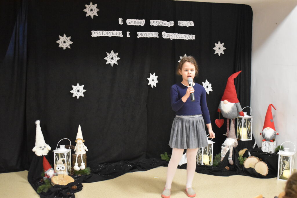 016 Stojąca dziewczynka trzyma w ręku mikrofon. W tle czarny materiał na którym widnieje napis II Gminny Przegląd Kolęd, Pastorałek i Piosenek świątecznych. Na podłodze stoją kolorowe skrzaty, lampiony i pieńki.