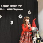 022 Dziewczynka w czerwonej sukience trzyma w ręku mikrofon. W tle czarny materiał na którym widnieje napis II Gminny Przegląd Kolęd, Pastorałek i Piosenek świątecznych. Na podłodze stoją kolorowe skrzaty, lampiony i pieńki.