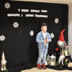 047 Dziewczynka w czarnej marynarce i białej sukience z warkoczami śpiewa do mikrofonu. W tle czarny materiał na którym widnieje napis II Gminny Przegląd Kolęd, Pastorałek i Piosenek świątecznych. Na podłodze stoją kolorowe skrzaty, lampiony i pieńki.