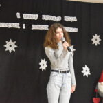 067 Mała dziewczynka w czerwonej spódnicy i białej bluzce trzyma mikrofon w ręku. W tle czarny materiał na którym widnieje napis II Gminny Przegląd Kolęd, Pastorałek i Piosenek świątecznych. Na podłodze stoją kolorowe skrzaty, lampiony i pieńki.