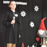 073 Nastolatka w szarej bluzce śpiewa do mikrofonu, w tle czarna kotara z białymi gwiazdkami i napisem, obok stoi krasnal w czerwonej czapce .