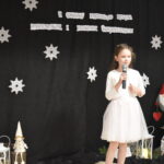 095 Chłopiec w przebraniu górala śpiewa do mikrofonu. W tle czarny materiał na którym widnieje napis II Gminny Przegląd Kolęd, Pastorałek i Piosenek świątecznych. Na podłodze stoją kolorowe skrzaty, lampiony i pieńki.