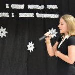 127 Panienka w czarnej sukience z białym kołnierzykiem trzyma mikrofon w ręku. w tle czarna kotara z białymi gwiazdkami i napisem za nią stoi skrzat.