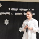 144 Dziewczynka w niebieskiej sukience śpiewa do mikrofonu. W tle czarny materiał na którym widnieje napis II Gminny Przegląd Kolęd, Pastorałek i Piosenek świątecznych. Na podłodze stoją kolorowe skrzaty, lampiony i pieńki.