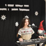 111 Dziewczynka trzyma w ręku mikrofon. W tle czarny materiał na którym widnieje napis II Gminny Przegląd Kolęd, Pastorałek i Piosenek świątecznych. Na podłodze stoją kolorowe skrzaty, lampiony i pieńki.