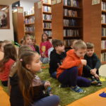 Przy drewnianym stole siedzą dzieci i wycinają z białego papieru. W tle regały z książkami, dwa okna i pomiędzy nimi duży zielony kwiat.