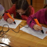 W bibliotece dzieci wycinają z kolorowych pianek wzory, na końcu stołu stoi kobieta i rozmawia z dziećmi w tle regały z książkami.