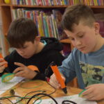 W bibliotece na zielonym dywanie siedzi kobieta i dzieci w kółku, za nimi regały z książkami.