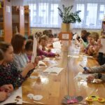 Dzieci siedzą przy drewnianym długim stole i wycinają nożyczkami szablony. Na stole są ścinki kartek. W oddali dwie kobiety rozdają kolorowe pianki. W tle pomieszczenia biblioteki z regałami pełnymi książek.