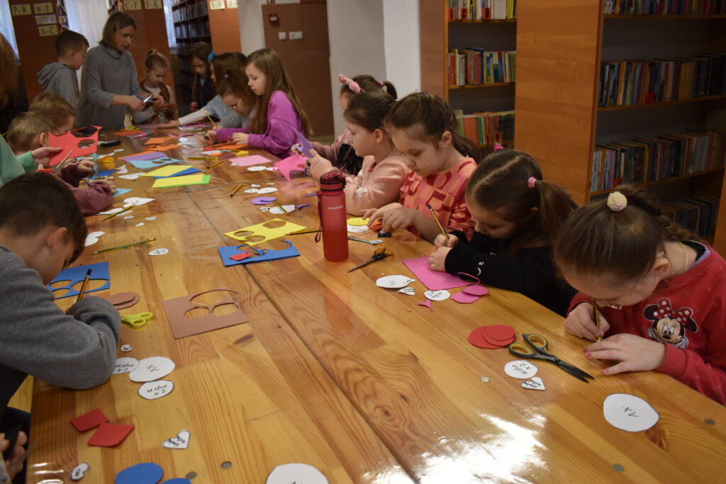 145 W bibliotece przy długim drewnianym stole na którym leżą nożyczki, ołówki i ścinki kolorowych kartek siedzą dzieci. Jedna dziewczynka podnosi rękę, jedna wycina. W tle dwa okna i regały z kolorowymi książkami.