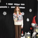 127 Panienka w czarnej sukience z białym kołnierzykiem trzyma mikrofon w ręku. w tle czarna kotara z białymi gwiazdkami i napisem za nią stoi skrzat.