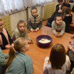 205 Przy długim drewnianym stole po obu stronach siedzą dzieci i malują przezroczyste szklanki. Przygląda im się kobieta w jasnej bluzce. W tle regały z książkami.