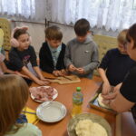 Chłopcy i dziewczęta w bibliotece siedzą przy długim drewnianym stole na którym leżą białe kartki i kredki. W tle stoi kobieta w pomarańczowym swetrze i trzyma w reku dużą żółta kostkę do gry za nią stoją regały z książkami.
