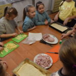 W bibliotece na zielonym dywanie siedzą dzieci w tle regały z książkami.