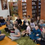 Dziewczynki i chłopcy siedzą przy stole i rysują pomarańczowymi długopisami w tle regały z książkami.