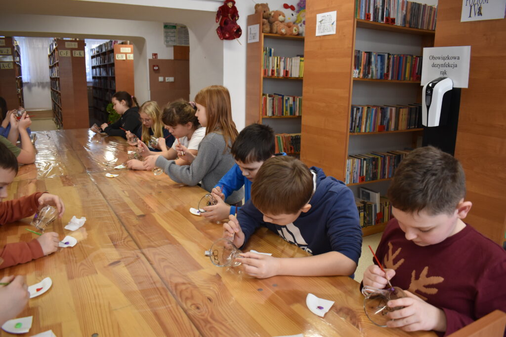 Dzieci siedzą przy stole na którym leżą kable i długopisy 3D w tle regały biblioteczne z książkami.