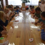 Dzieci siedzą i rysują pomaga im pani która stoi za nimi.