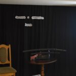 Mężczyzna w średnim wieku w niebieskiej koszuli i czarnej marynarce stoi przy jasnym krześle. W tle czarna kotara na której widnieje napis „Spotkanie z podróżnika” na stoliku stoją miecze samurajskie, z prawej strony stoi ekran