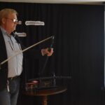 Mężczyzna w okularach, w niebieskiej koszuli i czarnej marynarce stoi i trzyma w ręku miecz. W tle czarna kotara i ekran.