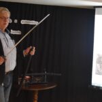 Stojący mężczyzna w średnim wieku trzyma w ręku miecz samurajski. W tle czarna kotara i ekran.