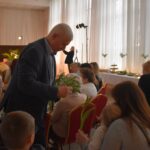 Na czerwonych krzesłach siedzą kobiety, stoi mężczyzna na ręku trzyma tulipany którymi obdarowuje panie. W tle stoły przykryte białymi obrusami na których stoją przekąski i wysokie wazony z kwiatami.