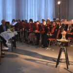 Na pierwszym planie mężczyzna w białej koszuli z kucykiem na głowie. W tle na czerwonych krzesłach siedzi orkiestra dęta w czerwono czarnych strojach.