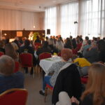 Na czerwonych krzesłach siedzą kobiety, stoi mężczyzna na ręku trzyma tulipany którymi obdarowuje panie. W tle stoły przykryte białymi obrusami na których stoją przekąski i wysokie wazony z kwiatami.