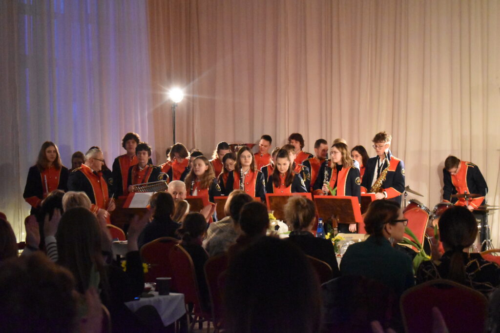 Orkiestra dęta gra na instrumentach przed nimi stoi dyrygent wszyscy są w czarno czerwonych mundurach i siedzą na czerwono złotych krzesłach. W oddali świeci reflektor.