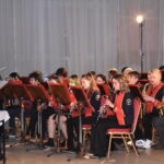 Orkiestra dęta gra na instrumentach przed nimi stoi dyrygent wszyscy są w czarno czerwonych mundurach i siedzą na czerwono złotych krzesłach. W oddali świeci reflektor.