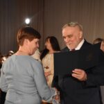 Trzy kobiety dostają po tulipanie od stojącego przed nimi siwiejącego mężczyzny. W tle duże okna z firanami.