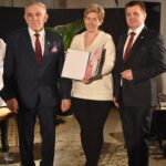 Mężczyzna w okularach, granatowym garniturze i czerwonym krawacie stoi trzymając w ręku ciemną teczkę.