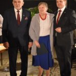 Kobieta w białej bluzce, niebieskiej spódnicy i czerwonych koralach. Obok niej stoi dwóch mężczyzn w czarnych garniturach