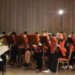 Orkiestra dęta gra na instrumentach przed nimi stoi dyrygent wszyscy są w czarno czerwonych mundurach i siedzą na czerwono złotych krzesłach. W oddali świeci reflektor.