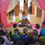 W pomieszczeniach biblioteki na środku siedzi kobieta po obu stronach są kolorowe kotary za nią regały z książkami, przed nią siedzą na zielonym dywanie dzieci. Kobieta trzyma w rękach przezroczystą kulę, obok niej stoi kolorowy kufer i leżą kolorowe poduszki.