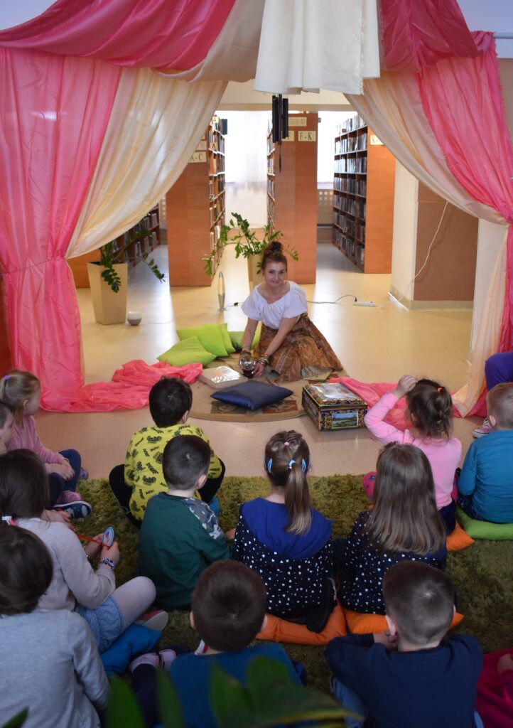 W pomieszczeniach biblioteki na środku siedzi kobieta po obu stronach są kolorowe kotary za nią regały z książkami, przed nią siedzą na zielonym dywanie dzieci. Kobieta trzyma w rękach przezroczystą kulę, obok niej stoi kolorowy kufer i leżą kolorowe poduszki.