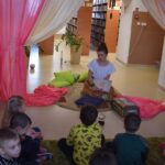 Siedząca kobieta na dywanie trzyma dużą książkę, po obu stronach różowo beżowe kotary, za nią regały z książkami i duży kwiatek w beżowej donicy. Przed nią siedzą dzieci po turecku