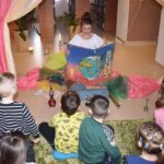 Kobieta w białej bluzce siedzi po turecku z dużym atlasem na którym widać planetę ziemię wokół niej porozkładane kolorowe materiały, przed nią siedzą dzieci. W tle regały z książkami i dwa kwiaty w beżowych donicach.