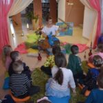 Na środku pomieszczenia siedzi kobieta w białej bluzce na dywani i ma w jednym ręku złotą lunetę w drugiej niebieską laleczkę.