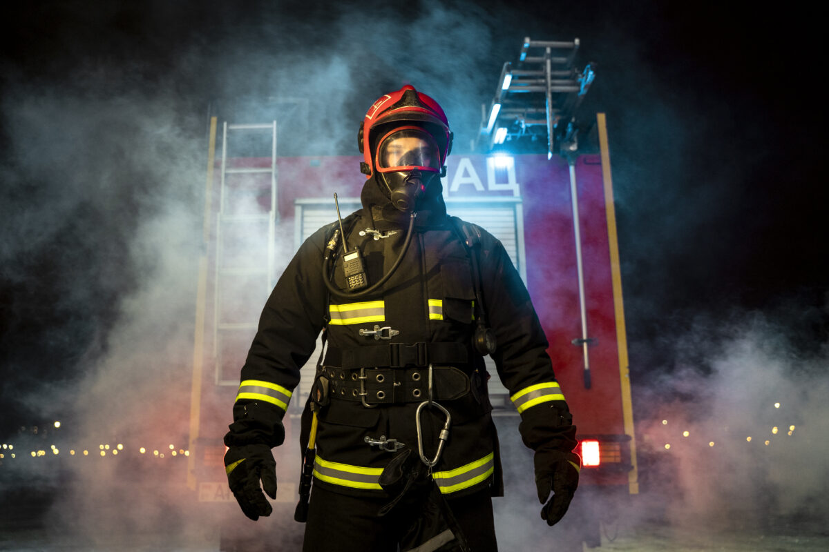 strażak w pełnym umundurowaniu stojący przed pojazdem strażackim