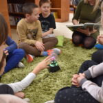 Na zielonym dywanie stoi kula a dzieci siedzące w kole dotykają ją rękoma