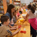Dzieci w Bibliotece na drewnianym stole zabierają się do pieczenia pierników. W tle regały z książkami