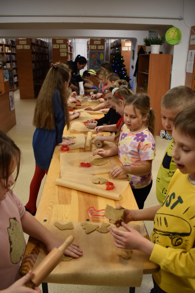 Dzieci w Bibliotece na drewnianym stole zabierają się do pieczenia pierników. W tle regały z książkami