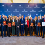 418435254_758076013016161_3825562095432386141_n Obraz przedstawia grupę osób stojących w dwóch rzędach, ubranych elegancko, większość mężczyzn w garniturach, a kobiety w sukienkach lub eleganckich garniturach. Na pierwszym planie sześć osób trzyma certyfikaty lub podobne dokumenty w brązowych okładkach. W tle stoi drugi rząd ludzi, również z dokumentami w rękach. Wszyscy patrzą w stronę kamery z uśmiechem i wyglądają na zadowolonych. Tło stanowi wnętrze o eleganckim charakterze, prawdopodobnie sala konferencyjna lub podobne pomieszczenie. Na ścianie w tle widnieje kilka flag, co może sugerować, że jest to wydarzenie o charakterze międzynarodowym lub rządowym. Pośrodku, na ścianie, znajduje się duże logo lub herb, co może wskazywać na konkretną instytucję lub organizację. Na podłodze jest wykładzina, a całe pomieszczenie jest dobrze oświetlone. Wydarzenie to wydaje się być ważną ceremonią lub uroczystością, być może wręczeniem nagród, certyfikatów lub uznania dla grupy ludzi. Atmosfera jest formalna i celebracyjna.