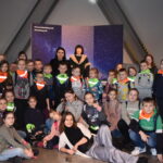 70 Duża grupa dzieci w odblaskowych chustach, pozuje do zdjęcia. W tle postać Mikołaja Kopernika