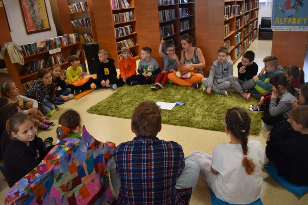90 W bibliotece na podłodze siedzą dzieci w kole w środku leży zielony dywan. W tle regały z książkami