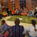 90 W bibliotece na podłodze siedzą dzieci w kole w środku leży zielony dywan. W tle regały z książkami