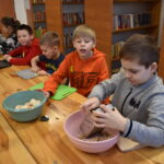 205 Przy stole siedzi czterech chłopców i dwie dziewczynki przed nimi leżą deski do krojenia i stoi miska z pieczarkami. Jeden chłopiec trze pieczarki na tarce. W tle regały z książkami.