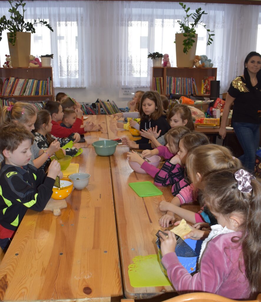 212 Dzieci siedzą przy stole przed nimi deski do krojenia, miski. W tle regały z książkami